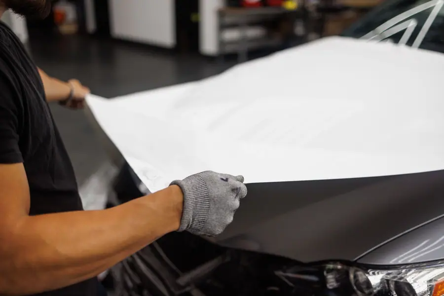 Installer in gloves stretching white vinyl wrap film over a car hood (stock photo)